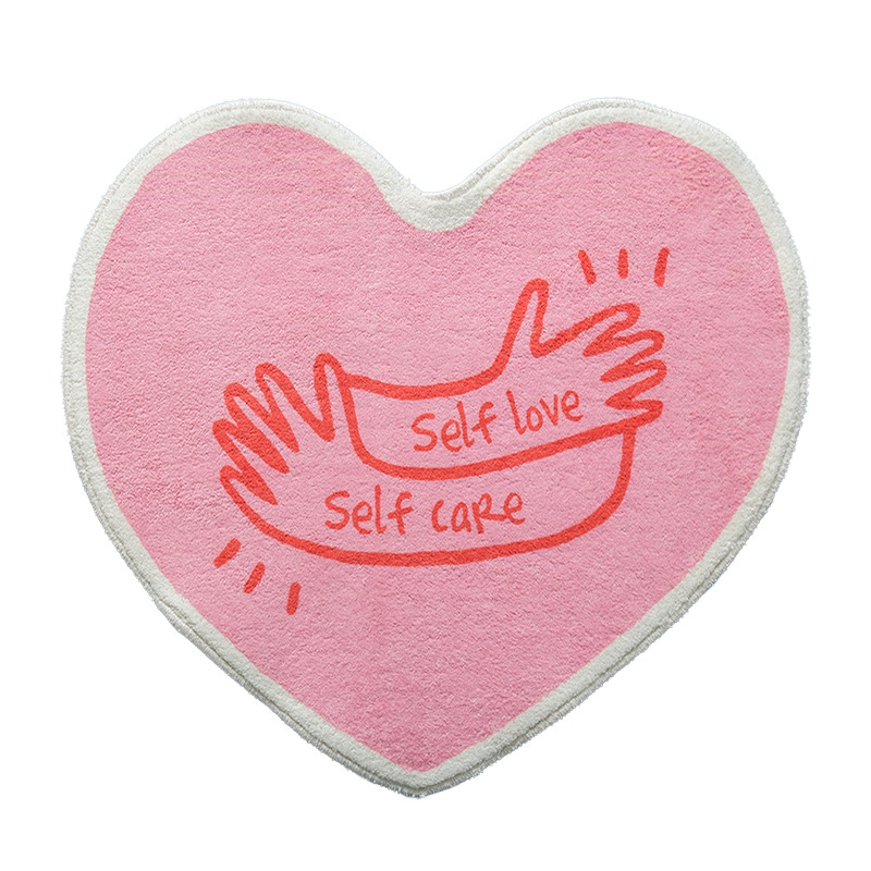 Alfombra de dormitorio para niñas en forma de corazón en color rosa. En la parte superior, un estampado de dos manos con las palabras self love self care en rojo.
