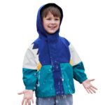 Un niño caucásico con una fina parka de patchwork con capucha verde, azul, blanca y amarilla sobre un fondo liso.