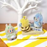 3 peluches Labubu con disfraces de animales de peluche, colocadas sobre una toalla amarilla y blanca, sobre una mesa de madera con un árbol blanco de fondo